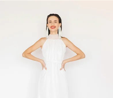 Eine lächelnde Frau in einem ärmellosen weißen Kleid steht vor hellem Hintergrund. Sie trägt auffällige lange Ohrringe und roten Lippenstift, während ihre Hände in die Taille gestützt sind.